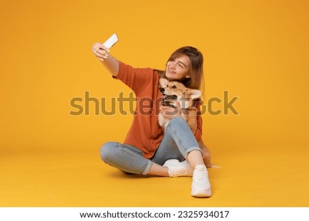 Excited european woman taking selfie with her pretty corgi dog, looking at phone camera and embracing pet, isolated on yellow studio background wall