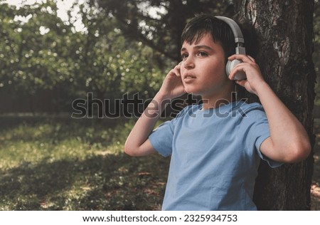 Toned portrait of a dark haired Caucasian adorable teenage boy, schoolkid in blue casual t-shirt, listening to music on wireless headphones, relaxing after class, sitting under a tree in the park Royalty-Free Stock Photo #2325934753
