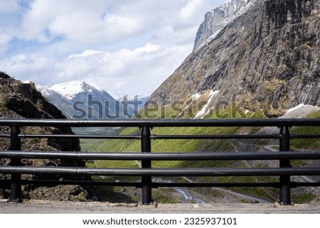 on an early spring day when there is snow on the mountain and the rocks are gray but above them is a blue sky with white clouds and there are metal railings on the side of the road