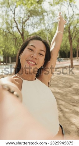 Smiling brunette girl dressed in white top and jeans, taking selfie on mobile phone while standing in city park