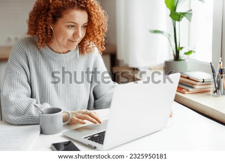 Portrait of curly haired redhead caucasian female in wired headphones spending leisure time using laptop during coffee break, sitting at kitchen table with smartphone, visiting popular web pages Royalty-Free Stock Photo #2325950181
