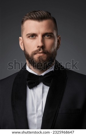 Close-up portrait of a stylish and attractive man with a well-groomed beard, dressed in a high-end black suit and bow tie, posing confidently on a gray background Royalty-Free Stock Photo #2325968647