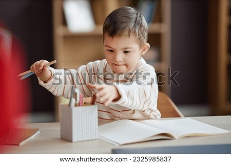 Portrait of excited little boy with down syndrome drawing pictures while sitting at table at home and using colored pencils 