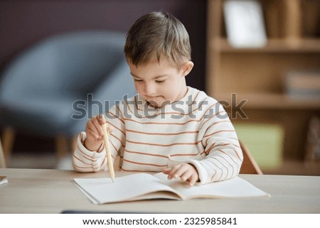 Portrait of cute little boy  with down syndrome drawing pictures while sitting at table at home and using pencils, copy space 
