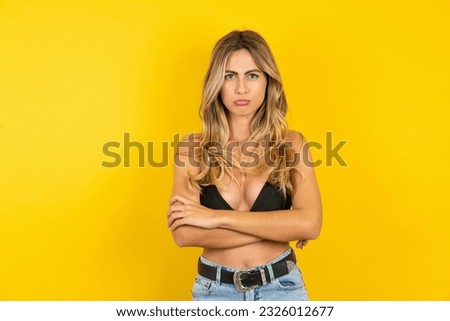 Gloomy dissatisfied Young blonde woman on vacation wearing bikini over yellow background looks with miserable expression at camera from under forehead, makes unhappy grimace