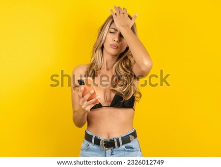 Upset depressed Young blonde woman on vacation wearing bikini over yellow background makes face palm as forgot about something important holds mobile phone expresses sorrow and regret blames Royalty-Free Stock Photo #2326012749