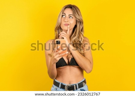 Young blonde woman on vacation wearing bikini over yellow background thinks deeply about something, uses modern mobile phone, tries to made up good message, keeps index finger near lips.