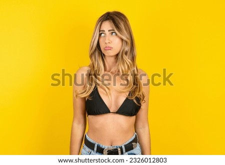 Dissatisfied Young blonde woman on vacation wearing bikini over yellow background purses lips and has unhappy expression looks away stands offended. Depressed frustrated model.