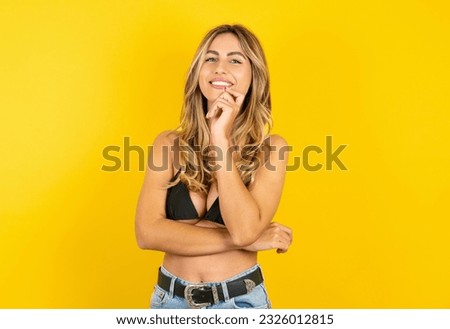 Young blonde woman on vacation wearing bikini over yellow background looking confident at the camera smiling with crossed arms and hand raised on chin. Thinking positive.