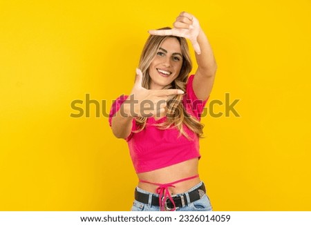 Young blonde woman wearing pink tank top yellow background making finger frame with hands. Creativity and photography concept.