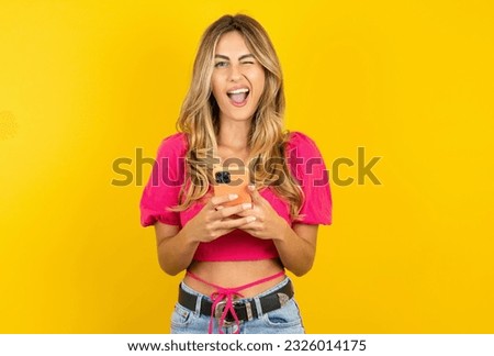 Excited Young blonde woman wearing tank top yellow background winking and eye hold smart phone use read social network news