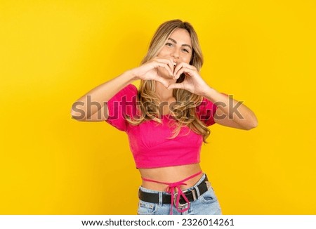 Young blonde woman wearing pink tank top yellow background smiling in love doing heart symbol shape with hands. Romantic concept.
