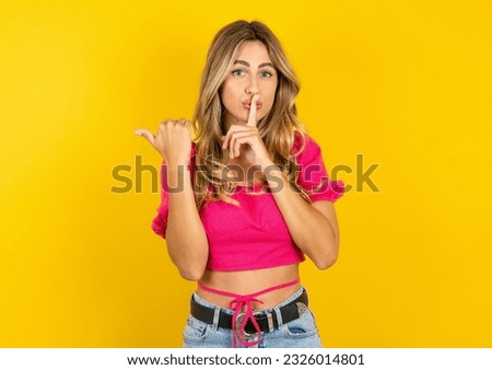 Young blonde woman wearing pink tank top yellow background asking to be quiet with finger on lips pointing with hand to the side. Silence and secret concept.