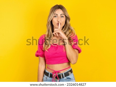 Young blonde woman wearing pink tank top yellow background makes silence gesture, keeps finger over lips. Silence and secret concept.
