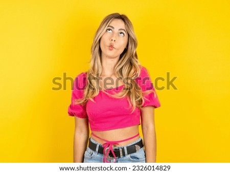 Young blonde woman wearing pink tank top yellow background making fish face with lips, crazy and comical gesture. Funny expression.