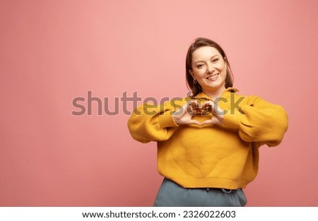 Adult woman smiling, showing hand heart gesture over chest and staring at camera, making love confession, standing against pink background. Royalty-Free Stock Photo #2326022603