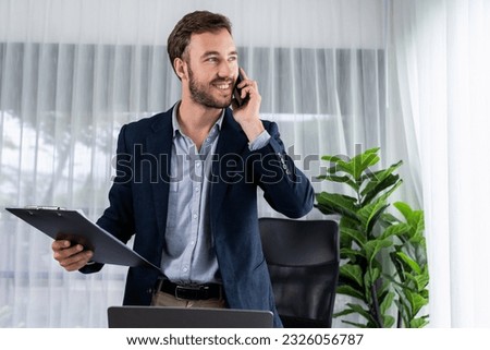Hardworking businessman stand confidently in modern office making persuasive sales call to client. Office worker talking on the phone coordinate and manage business work with colleagues. Entity Royalty-Free Stock Photo #2326056787