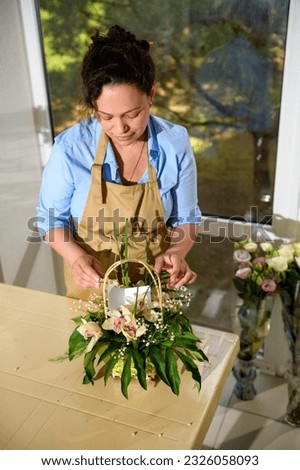 Overhead view of a female florist creating a flower arrangement with fresh orchid flowers and plants, inserting a birthday card for special festive life event, with copy space, in floral design studio