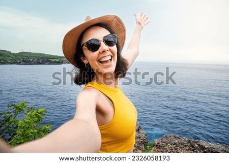 Young woman traveler wearing hat and sunglasses taking selfie in Nusa Penida, Bali. Female taking picture in vacation.