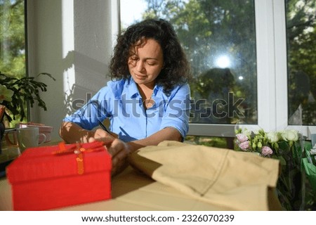 Beautiful curly haired Latin American young woman taking notes in her floral workshop. A beige apron and a red gift box on the table. People. Hobby. Small business. Lifestyle. Florist. Floriculturist. Royalty-Free Stock Photo #2326070239