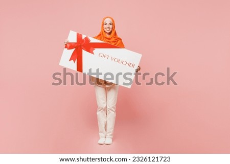 Full body young arabian asian muslim woman wear orange abaya hijab hold big gift certificate coupon voucher card for store isolated on plain pink background. Uae middle eastern islam religious concept
