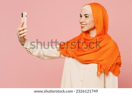 Young fun arabian asian muslim woman wear orange abaya hijab doing selfie shot on mobile cell phone isolated on plain light pink background studio portrait. Uae middle eastern islam religious concept