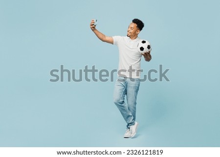 Full body young man fan wear t-shirt cheer up support football sport team hold in hand soccer ball watch tv live stream doing selfie shot on mobile cell phone isolated on plain blue color background