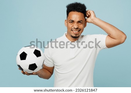 Young displeased disappointed man fan wear basic t-shirt scratch head cheer up support football sport team hold in hand soccer ball watch tv live stream isolated on plain pastel blue color background Royalty-Free Stock Photo #2326121847