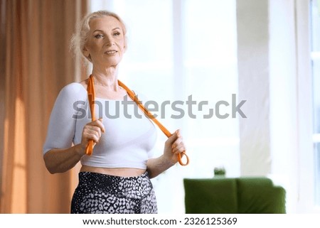Beautiful senior woman in comfortable sportswear standing with fitness equipment for home training in living room. Concept of sportive and healthy lifestyle, age, body care, fitness, wellness.