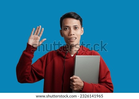 Excited handsome young Asian man wearing red hoodie greeting and greeting with hand while looking at someone isolated on blue background.