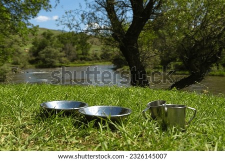Close-up of aluminum plates and mugs stand on the green grass against the incredible landscape of a mountain river and trees on a salty day. Cooking while traveling by car.