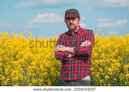 Portrait of confident farmer in blooming canola rapeseed field. Farm worker wearing red shirt and brown trucker's hat at plantation. Selective focus. Royalty-Free Stock Photo #2326151037