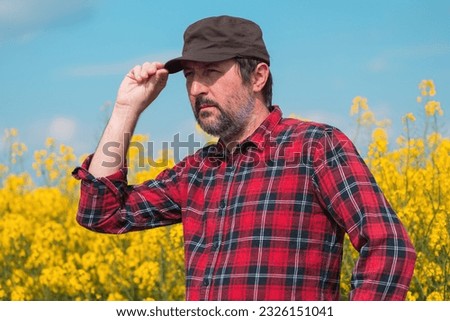 Portrait of confident farmer in blooming rapeseed field. Farm worker wearing red shirt and brown trucker's hat at plantation. Selective focus. Royalty-Free Stock Photo #2326151041
