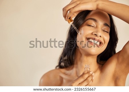 Happy young woman with bare shoulder applying serum on face with closed eyes. Beautiful hispanic young woman moisturizes her skin with serum isolated against beige background with copy space. Royalty-Free Stock Photo #2326155667