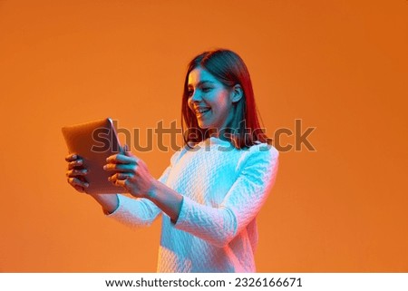 Portrait of young girl, student working on laptop, using gadgets against orange studio background in neon light. Concept of human emotions, youth, feelings, fashion, lifestyle, ad