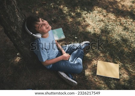 View from above of a Caucasian schoolboy in headphones, relaxing sitting under the tree in the park, smiling, enjoying cool soundtrack, listening to music after classes. People. Lifestyle. Education Royalty-Free Stock Photo #2326217549