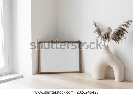 Dry grass, reed plant bouquet in modern ceramic vase. Empty photo frame mockup against white wall on wooden table near window. Minimalistic Scandinavian style, elegant boho interior, living room. 