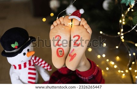 bare feet of a child on which the numbers 2024 are written, luminous garlands, a toy snowman. Cozy, festive Christmas atmosphere. Santa's helper. Cheerful childhood. selective focus Royalty-Free Stock Photo #2326265447