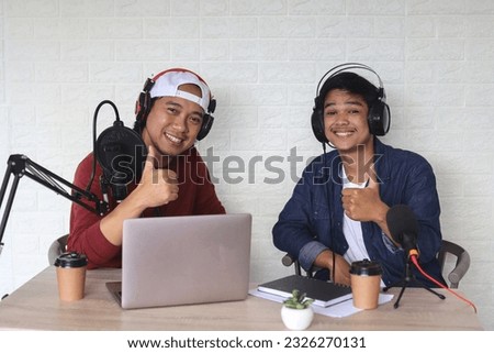 Two cheerful radio hosts or podcasters, smiling at camera and showing thumbs up while getting ready for a podcast live show in studio. 