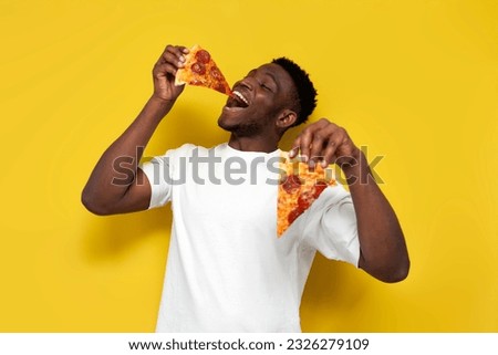 joyful african american man in white t-shirt eats piece of pizza and offers you on yellow isolated background, the guy gives and advertises fast food Royalty-Free Stock Photo #2326279109