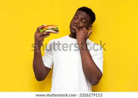 african american man in white t-shirt holds big burger and licks the sauce from his finger on yellow isolated background, young guy eats fast food, unhealthy eating concept Royalty-Free Stock Photo #2326279113