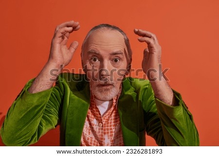 astonished senior man with bulging eyes holding hands near head and looking at camera on red orange background, trendy model, grey hair, beard, fashionable aging concept