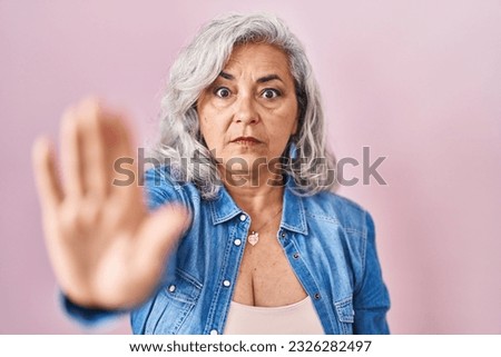 Middle age woman with grey hair standing over pink background doing stop sing with palm of the hand. warning expression with negative and serious gesture on the face. 