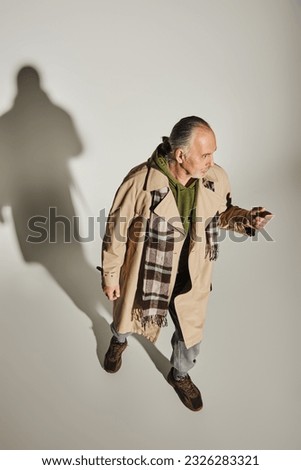 full length of senior man in stylish casual attire standing on grey background with shadow and looking away, green hoodie, beige trench coat, plaid scarf, fashionable aging concept, high angle view