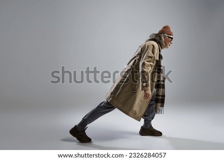 side view of senior and trendy model in hipster style outfit and dark sunglasses stepping on grey background with copy space, beanie hat, beige trench coat, sneakers, full length view, fashion shoot Royalty-Free Stock Photo #2326284057