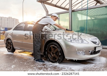 young guy in uniform car wash worker washes car with sponge with foam, man in overalls works in outdoors car cleaning service