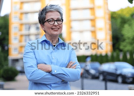 Smiling senior woman realtor crossed her arms over her chest against the backdrop of a high-rise building.
