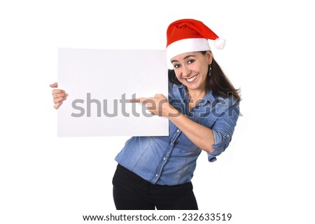 young attractive Hispanic woman wearing Santa Claus Christmas hat and blue shirt pointing blank billboard or placard sign as copy space for adding corportate marketing isolated on white background