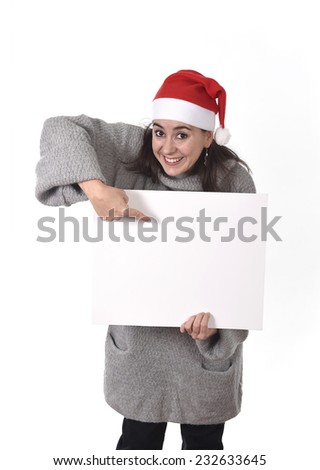 young attractive Hispanic woman wearing Santa Claus Christmas hat and winter jumper holding pointing blank billboard placard sign as copy space adding corporate marketing isolated on white background