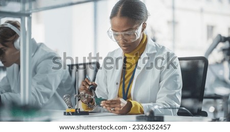 Diverse Team of Multiethnic Young Scientists Passing Internship in a Modern High Tech Laboratory. African Female Working with Soldering Iron Wearing Safety Goggles and White Lab Coat Royalty-Free Stock Photo #2326352457